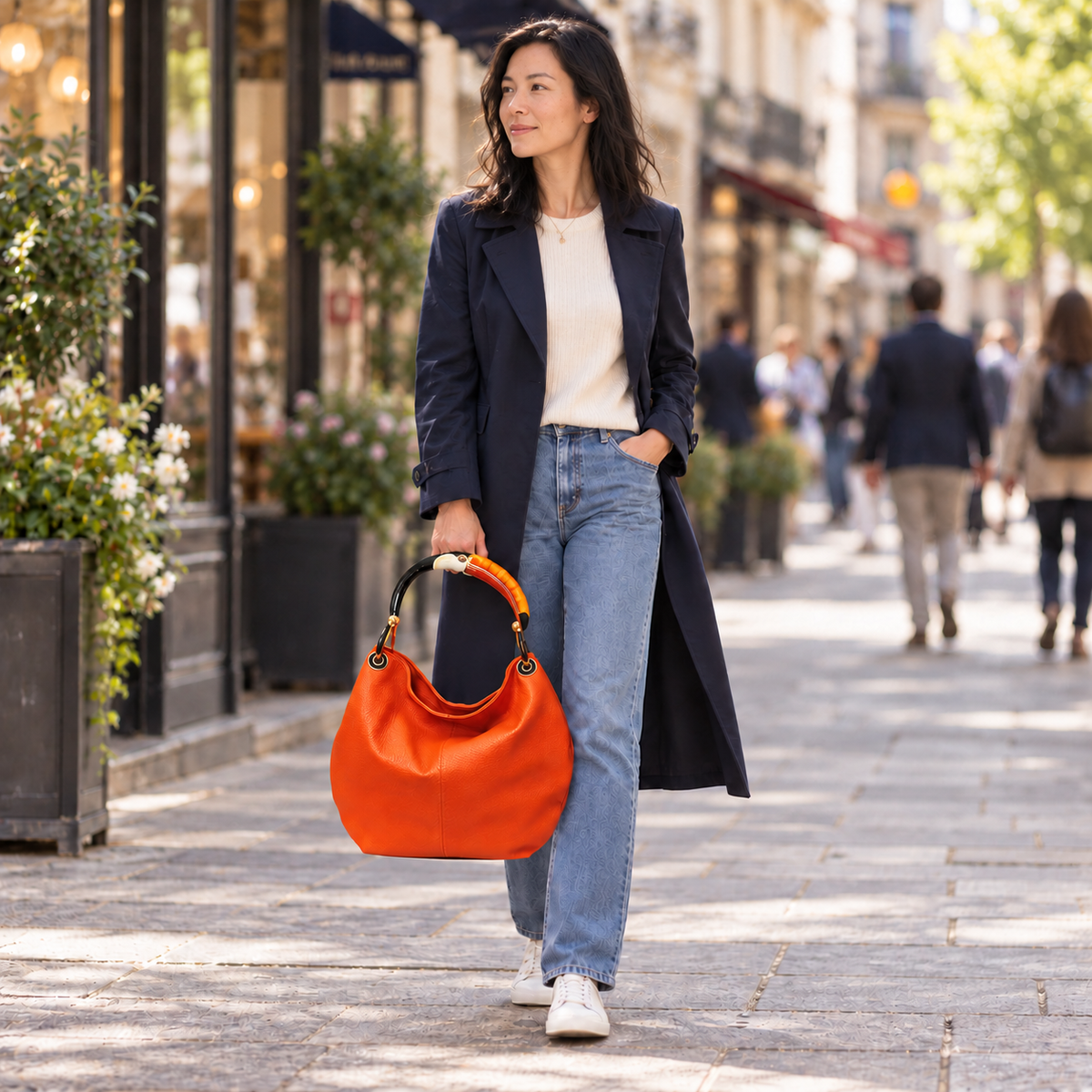 Woman holding a large orange leather bag with toucan sculpture handle walking along a city sidewalk in a navy coat and jeans.